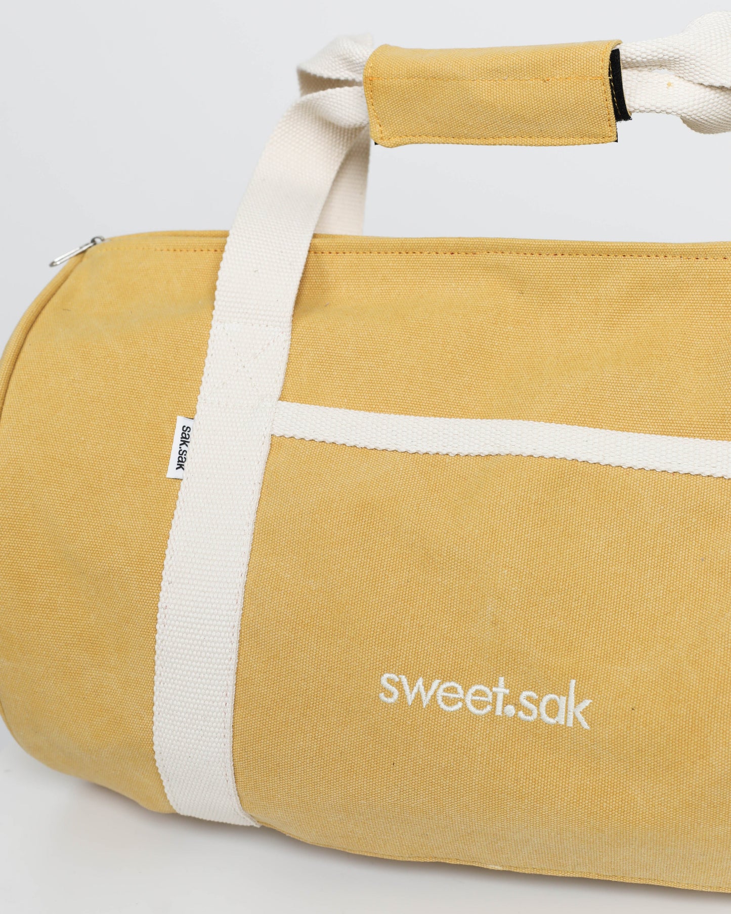 Yellow duffel bag with white straps and 'sweet.sak' branding on a light gray background