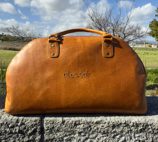 Brown leather duffel bag with 'alles sak' branding on a stone surface with a grassy field and trees in the background.