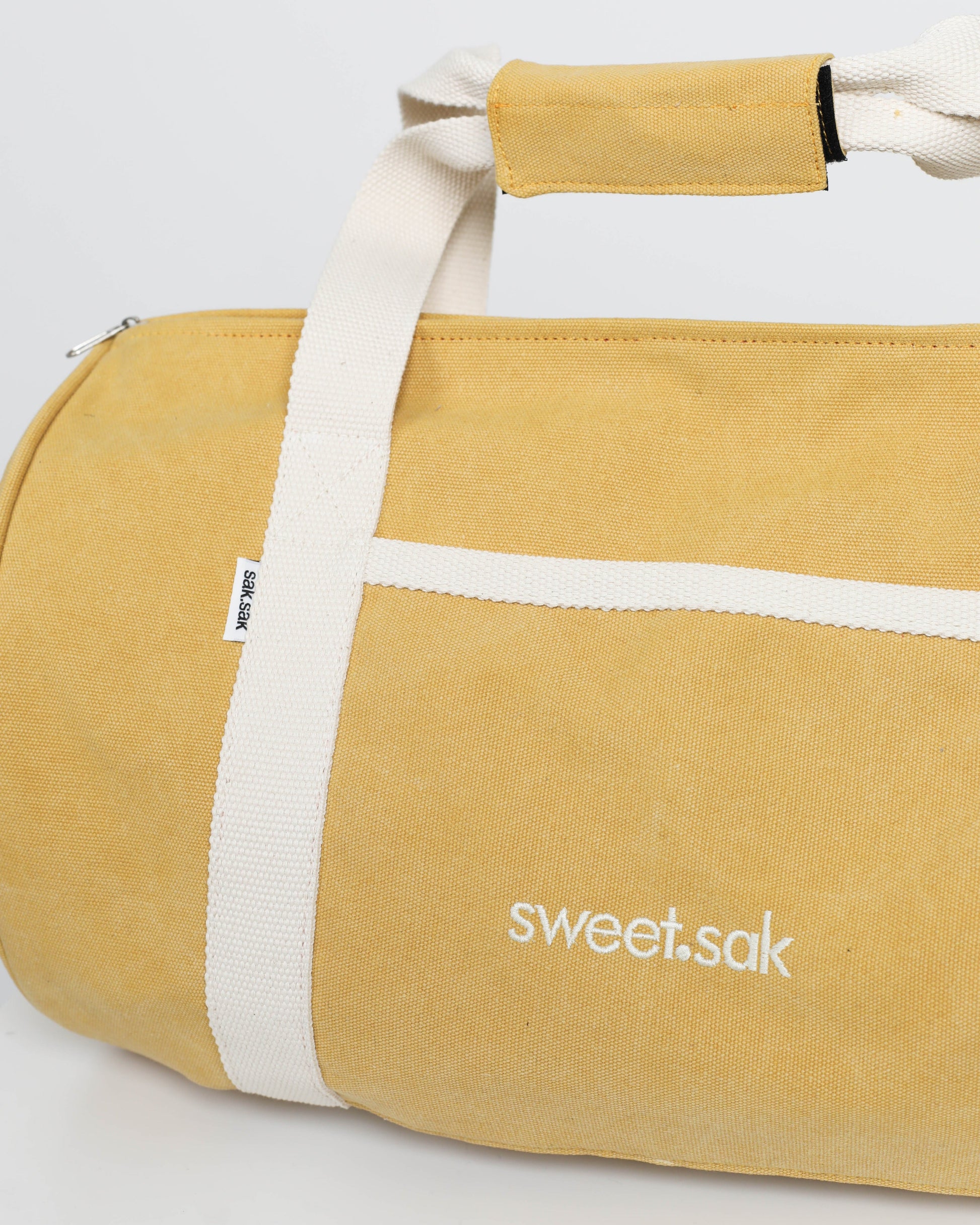 Yellow duffel bag with white straps and 'sweet.sak' branding on a light gray background