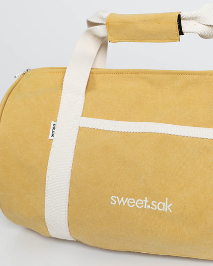 Yellow duffel bag with white straps and 'sweet.sak' branding on a light gray background